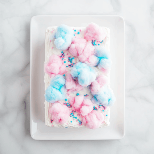 Top-down view of the chilled cake in the pan, now topped with a smooth layer of whipped topping and decorated with pink and blue cotton candy. The pan is placed over a white marble cooktop. Ingredients in the scene: whipped topping, cotton candy, chilled cake. Equipment: baking pan. The background remains clean and minimal --ar 4:6