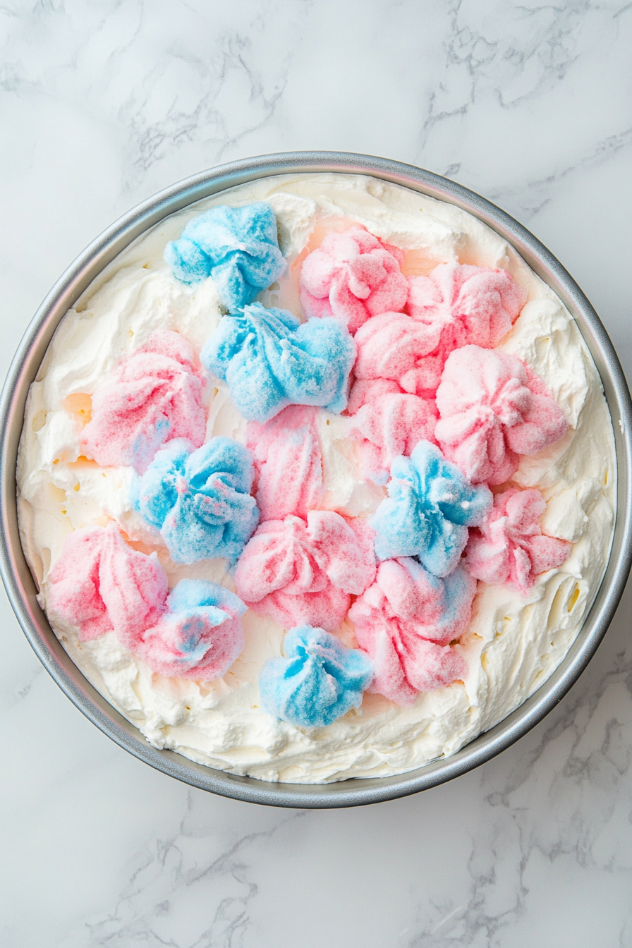 Top-down view of the fully prepared and decorated cotton candy cake placed on a white ceramic cake stand over a white marble countertop. The cake is topped generously with pastel pink and blue cotton candy and whipped topping. It looks whimsical, fluffy, and festive, ready to be served as a dreamy dessert centerpiece.
