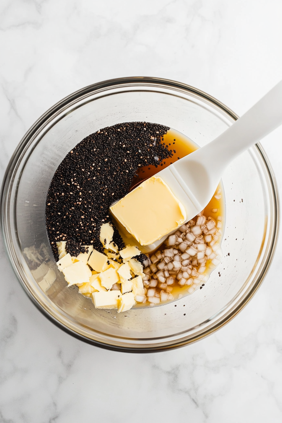 Top-down shot of a large clear glass bowl filled with melted butter, Dijon mustard, Worcestershire sauce, poppy seeds, and dried minced onion. A white silicone spatula rests inside, ready to mix. This flavorful mixture forms the glaze that will be brushed over the sandwiches later. The scene is set on a white marble countertop for a clean look.