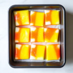 Top-down view of the Candy Corn Fudge after being sliced into small squares, showing its distinct yellow, orange, and white layers stacked atop one another. The fudge is arranged on a white dish, ready to serve on a white countertop.