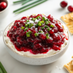 The strained cranberry mixture layered over the cream cheese base on a serving platter, garnished with green onion greens, flaky sea salt, and a drizzle of honey, accompanied by crackers on the white marble cooktop.