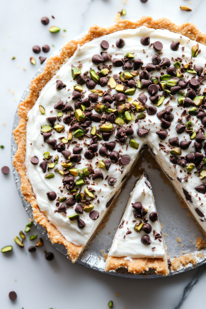 A chilled cannoli cream pie topped with mini chocolate chips and chopped pistachios, neatly sliced with a glossy finish, displayed on a clean white marble background.