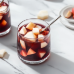 Glasses filled with chilled Peruvian Chicha Morada on the white marble countertop, garnished with floating apple pieces and served with a straw and spoon.