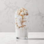 A tall glass filled with ice cubes and freshly poured oat milk, topped with a decorative drizzle of maple syrup, resting on a spotless white marble counter.