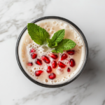 A completed cocktail in a Collins glass, topped with fizzy seltzer, garnished with a sprig of mint and vibrant pomegranate seeds, set on the white marble cooktop.