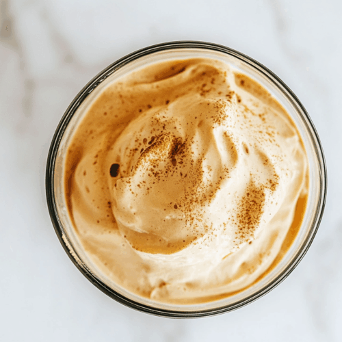 A top-down view of the finished Dalgona Coffee served in a transparent glass placed on a white marble countertop. The glass shows a creamy layer of whipped coffee sitting perfectly on top of a base of cold milk and ice cubes. The clean and minimal setup emphasizes the contrast between the fluffy brown coffee and the milky white base. This visually appealing beverage is known for its frothy texture and rich aroma, created from just three simple ingredients: instant coffee, sugar, and water, whipped until airy and spooned over chilled milk. It's a trending and delicious drink.