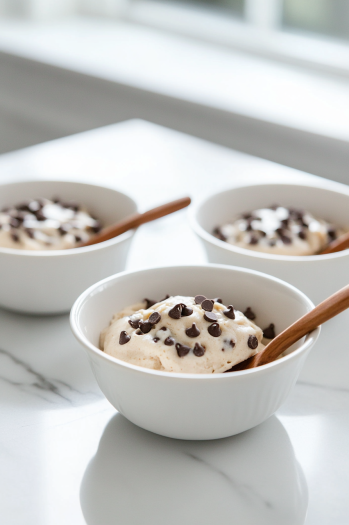 Spoonfuls of edible cookie dough served in small bowls on the white marble cooktop, with chocolate chips visible throughout the dough, ready to be enjoyed directly from the bowl.