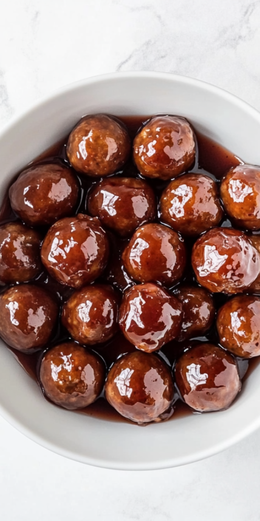 Top-down view of sweet and savory meatballs cooked in a slow cooker using frozen meatballs, ketchup, and grape jelly. The meatballs are served in a white bowl, glistening with thick, tangy red-brown sauce. The image highlights the finished dish on a white marble countertop, showcasing the richness and appeal of this classic party appetizer.