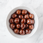Top-down view of sweet and savory meatballs cooked in a slow cooker using frozen meatballs, ketchup, and grape jelly. The meatballs are served in a white bowl, glistening with thick, tangy red-brown sauce. The image highlights the finished dish on a white marble countertop, showcasing the richness and appeal of this classic party appetizer.