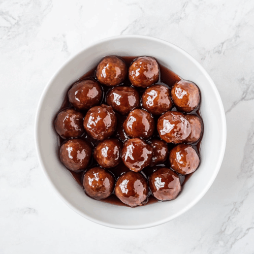Top-down view of sweet and savory meatballs cooked in a slow cooker using frozen meatballs, ketchup, and grape jelly. The meatballs are served in a white bowl, glistening with thick, tangy red-brown sauce. The image highlights the finished dish on a white marble countertop, showcasing the richness and appeal of this classic party appetizer.