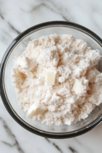 Close-up shot of a glass mixing bowl on the white marble cooktop, filled with all-purpose flour, salt, and sugar. Cold, cubed vegan butter is scattered throughout the mixture, ready to be cut in.