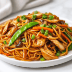 side view of a beautifully plated dish of stir-fried chicken with Hong Kong-style noodles, garnished with vibrant snap peas, carrots, mushrooms, mung bean sprouts, and scallions. The noodles are golden and slightly crispy, glistening with soy sauce and sesame oil. The clean and simple presentation over a white marble countertop adds elegance to the vibrant and fresh-looking meal, ready to be served with cucumber salad or grapefruit crush.