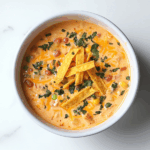 A comforting bowl of creamy chicken taco soup served in a white ceramic bowl placed on a clean white marble countertop. The soup is topped with golden corn tortilla strips, melted cheddar cheese, and visible chunks of chicken, corn, beans, and tomatoes. The overall look is hearty, vibrant, and full of rich Mexican-inspired flavors, making it an inviting dish for any mealtime.