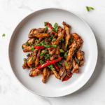 A delicious plate of crispy chilli chicken coated in a glossy, sticky sauce made from soy sauce, honey, and sriracha. The chicken is garnished with fresh chopped spring onions and served on a white ceramic plate placed on a clean white marble countertop, capturing the rich texture and mouthwatering appeal of this Indo-Chinese dish.