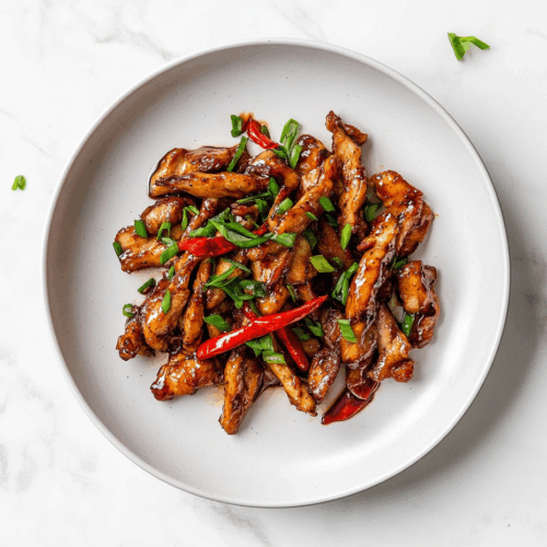 A delicious plate of crispy chilli chicken coated in a glossy, sticky sauce made from soy sauce, honey, and sriracha. The chicken is garnished with fresh chopped spring onions and served on a white ceramic plate placed on a clean white marble countertop, capturing the rich texture and mouthwatering appeal of this Indo-Chinese dish.