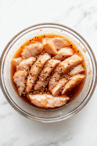 Raw chicken breast strips are shown marinating in a mixture of dark soy sauce, garlic and ginger paste, sesame oil, sugar, and white pepper. The marination is being done in a transparent glass bowl placed on a clean white marble countertop. This step ensures the chicken absorbs all the rich umami and aromatic flavors before coating and cooking.