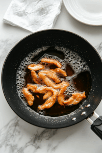 Chicken strips are being shallow fried in a black non-stick frying pan filled with hot vegetable oil. The chicken is golden brown and crispy, resting just above the bubbling oil. This step is essential for locking in flavor and creating the crispy exterior. A paper towel-lined plate is visible for draining excess oil.