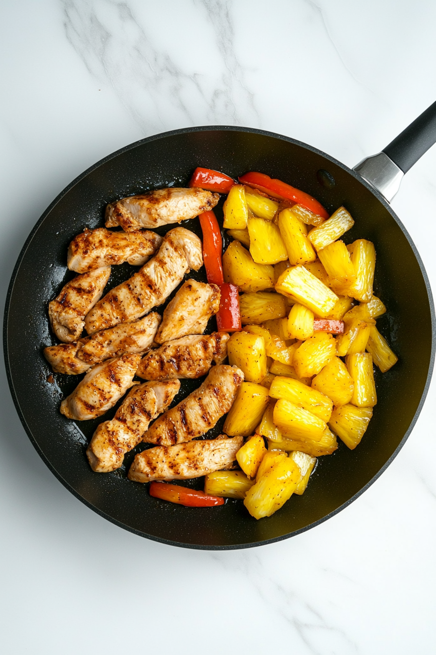 Top-down view of cooked chicken strips and bright yellow pineapple chunks added to sautéed red bell peppers and scallions in a black skillet. The white marble countertop provides a clean and minimal backdrop. The mix of juicy chicken and sweet pineapple creates a colorful blend of textures and flavors. Ingredient bowls for pineapple and reserved chicken are still visible in the scene, showcasing the layering process before the sauce is added to complete the dish.
