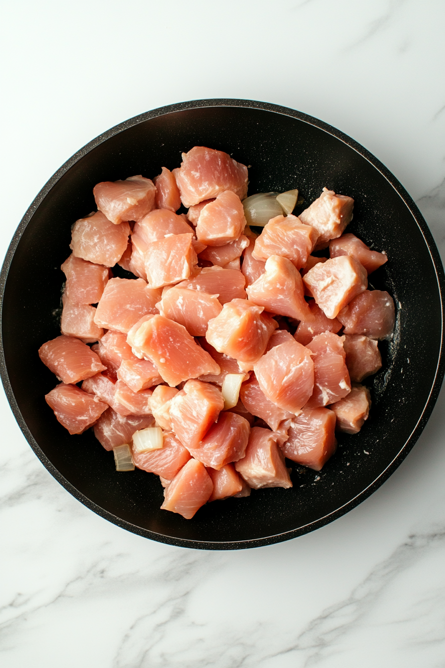 Top-down view of bite-sized chunks of boneless skinless chicken breast added to a black skillet already containing golden onions and garlic. The chicken is beginning to brown slightly on all sides. This essential step sets up the protein base of the butter chicken, with the white marble countertop providing a clean, professional backdrop to the process.