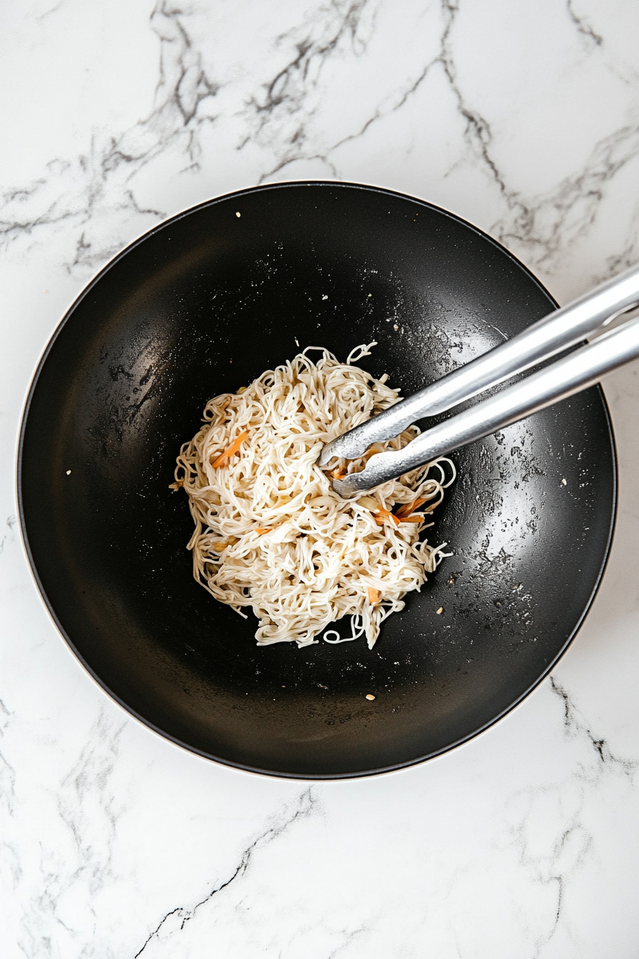 Top-down image of thin Hong Kong-style pan-fried noodles being added into the black wok already filled with sautéed vegetables. A blend of light soy sauce, dark soy sauce, sesame oil, and oyster sauce is being poured in, coating the noodles. Stainless steel tongs are shown tossing the noodles and mixing them with the vegetables. All of this is done on a white marble cooktop, highlighting the richness and balance of the dish.