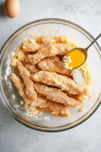 The marinated chicken is being coated with cornflour and egg white along with an extra spoon of garlic and ginger paste. This step is captured in a clear glass mixing bowl over a white marble countertop. This coating gives the chicken a light, crispy texture when fried and also helps the sauce cling beautifully in later steps.