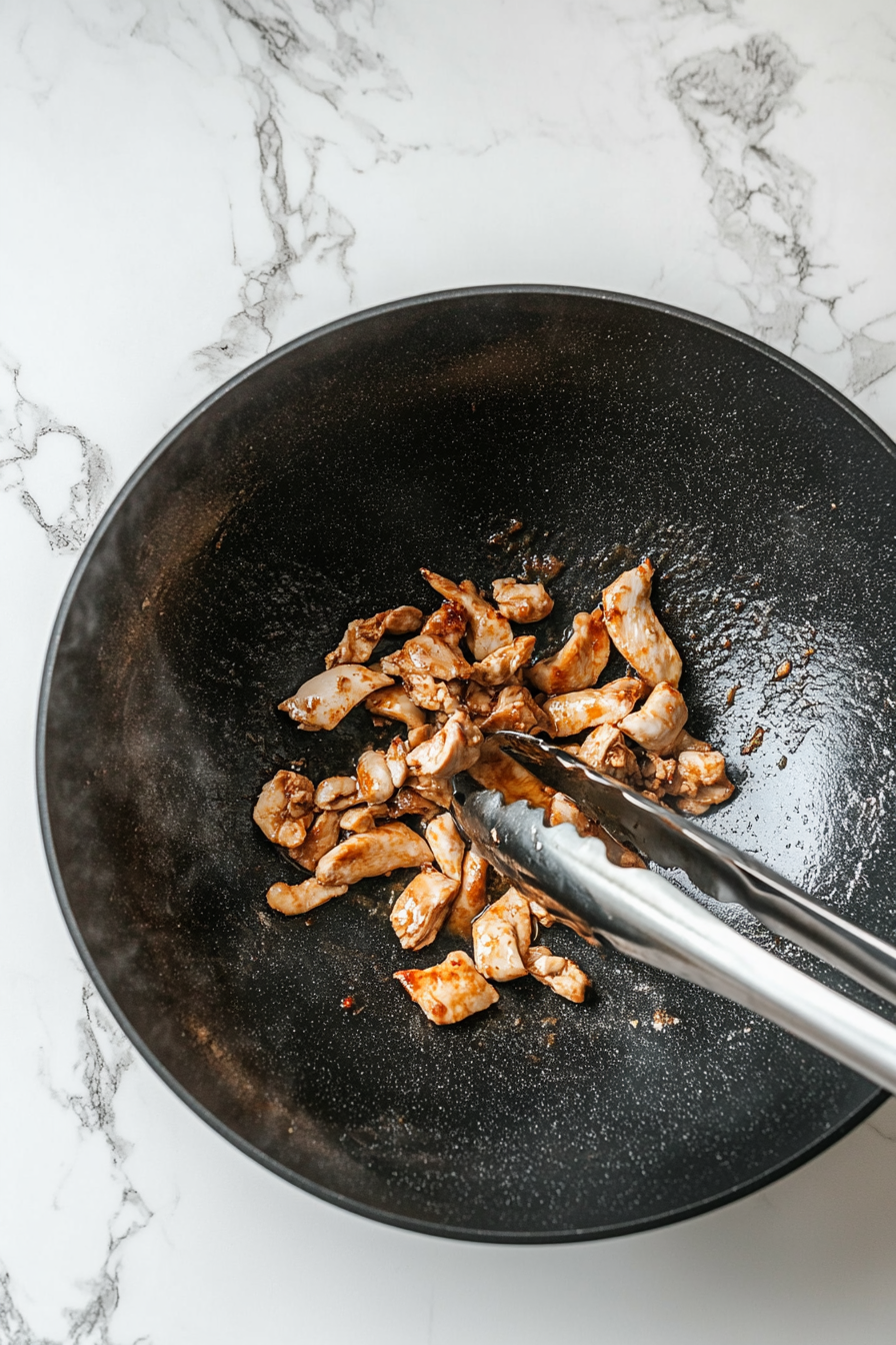 Top-down image of thinly sliced marinated chicken breast cooking in a black wok placed over a clean white marble cooktop. The chicken is turning light golden as it sizzles in hot oil, stirred with stainless steel tongs. Some marinade rests in a bowl beside the wok. This step is crucial to ensure the chicken is tender and juicy before being set aside for later mixing with vegetables and noodles.