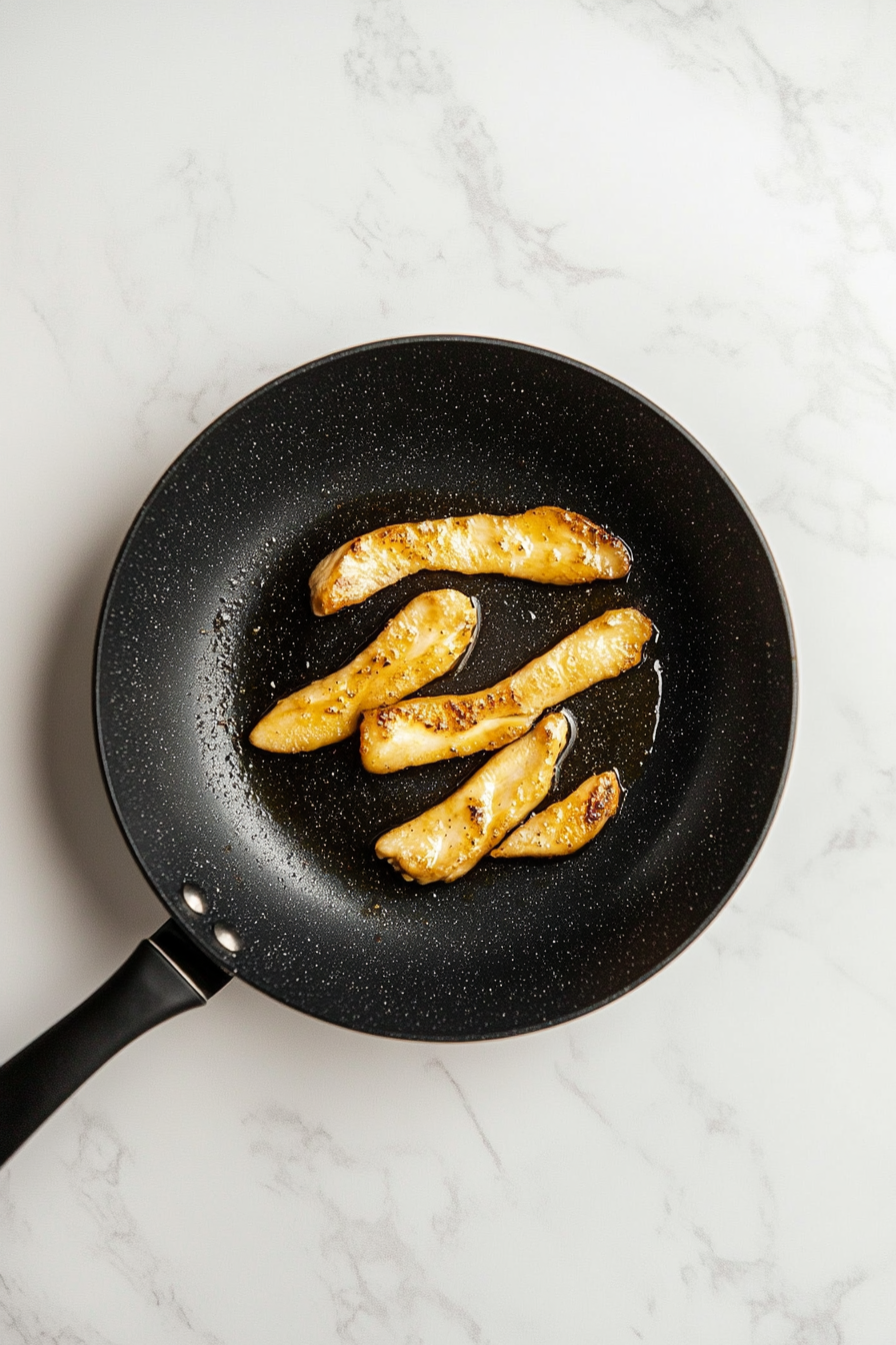 Top-down shot of golden brown chicken strips cooking in a black non-stick skillet placed on a clean white marble countertop. The marinated chicken is sizzling, developing a caramelized surface as it cooks through. A wooden spatula lies next to the skillet, and nearby small bowls show remnants of soy sauce and olive oil. The image captures the transformation of raw marinated chicken into beautifully seared strips full of flavor and texture.