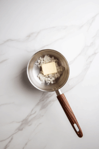 A stainless steel saucepan over a white marble surface contains melted butter and minced onion being sautéed with a wooden spatula. The onions are beginning to soften and turn translucent. A glass bowl with more butter and another with minced shallots sit nearby, showing the ingredients in progress as they build flavor for the creamy filling.