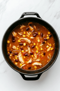 The soup mixture is gently simmering in a black pot with a partially covered lid on a white marble cooktop. Chicken breasts are slowly cooking while the soup bubbles lightly. Visible are beans, tomatoes, corn, and a golden broth enriched with spices. This step ensures tender chicken and flavor development, with the background kept clean and distraction-free for focus on the cooking process.