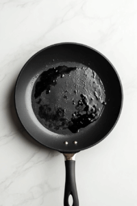 A mixture of dark soy sauce, honey, sriracha, and lemon juice is being simmered in a black non-stick skillet over a white marble countertop. The sauce is slightly thickening with small bubbles forming, indicating it is nearly ready. This bold and tangy stir fry sauce adds the signature flavor to the crispy chilli chicken.