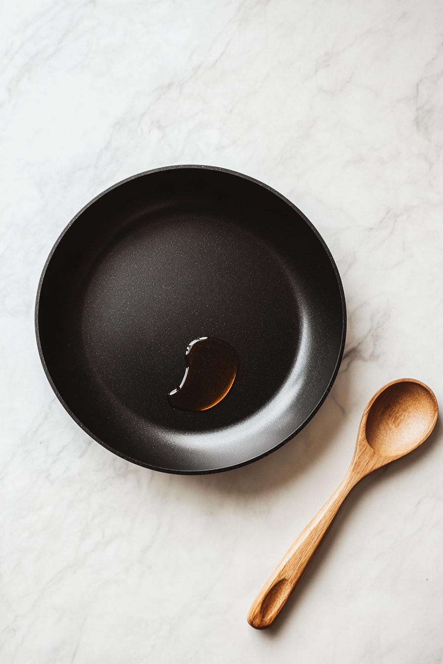 One tablespoon of vegetable oil is heating in a large black non-stick skillet placed on a clean white marble countertop. The oil is shimmering, indicating it's ready to cook the seasoned chicken. This step ensures the pan is properly preheated for searing the chicken to golden perfection. The setup is clean, simple, and focused.