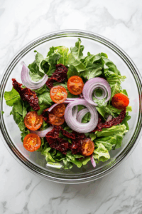 In a bowl, combine baby salad leaves, sliced red onion, and drained semi-dried tomatoes.