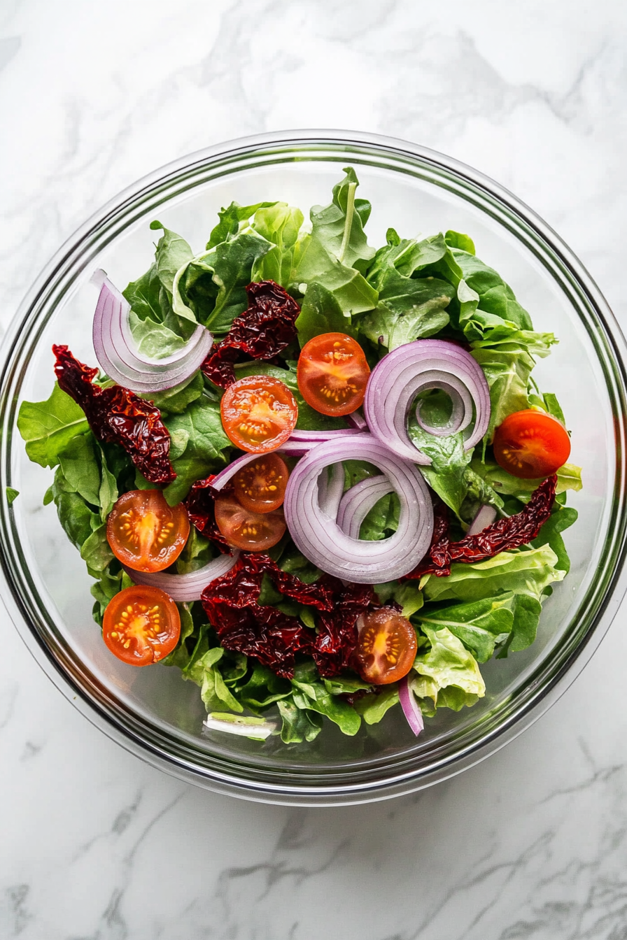 In a bowl, combine baby salad leaves, sliced red onion, and drained semi-dried tomatoes.